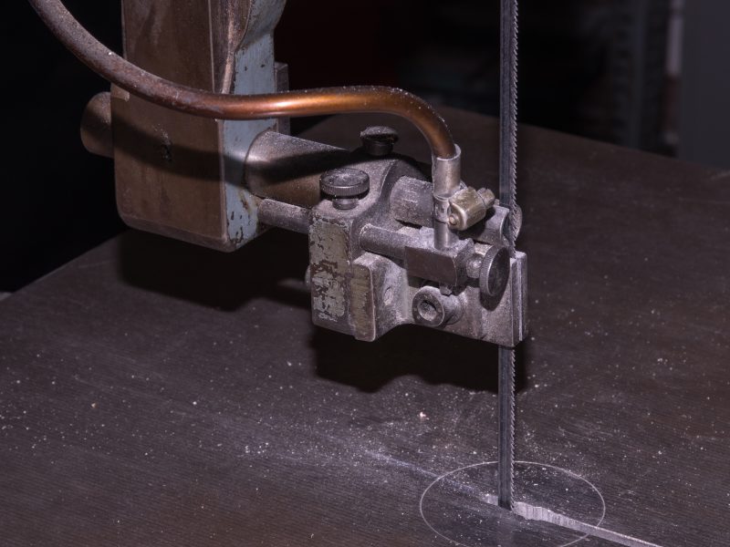 Close-up of a metal band saw in operation, with the blade cutting through a dark metal sheet in an industrial workshop.